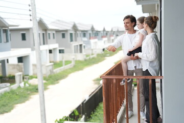 Fototapeta premium Young parents holding their child stand at the wooden railing admiring the neighborhood view on the balcony of their new house overlooking the residential estate in warm natural light