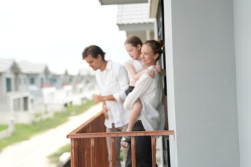 Fototapeta premium Happy young family with little daughter stand at the wooden railing admiring the neighborhood view standing on the terrace of a newly built suburban home on a bright sunny day