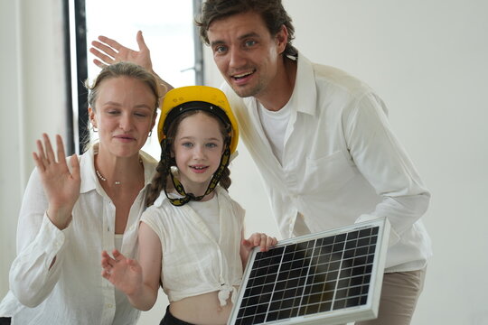 Young parents teaching their curious child kiss their daughter who proudly holds the photovoltaic module in the bright interior of their newly built home, shallow depth of field