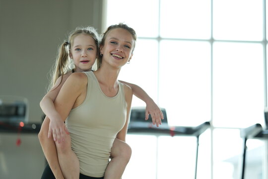 Loving mom and her cute child play piggyback, the girl hugging mom from behind after a family workout in the gym with copy space. Concept of family love, care and quality time