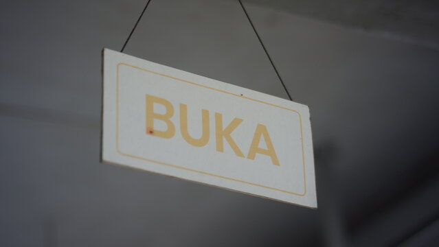 Buka sign, a hanging white rectangular wooden board with light orange text, indicating an open status for business, retail, or service establishments, a welcoming message for customers.