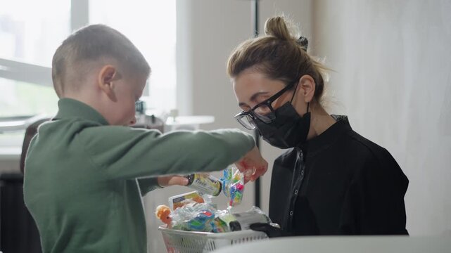 Young boy places items into basket while interacting with adult woman in soft lighting. scene takes place indoors featuring close up details of hands and objects during exchange. Both people