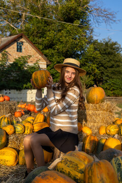 Young woman in straw hat holding a pumpkin in a sunny autumn garden, cheerful seasonal portrait surrounded by colorful gourds and rustic hay bales.