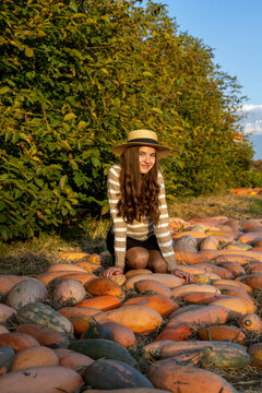 Young woman in straw hat posing among many pumpkins in a sunny autumn field, warm golden light creates a rustic seasonal harvest atmosphere.
