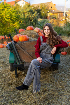 Young woman in denim overalls posing beside a rustic cart filled with pumpkins and hay, cozy autumn harvest portrait in warm evening light.
