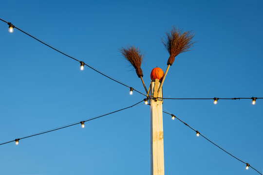 Rustic Halloween decoration with pumpkin head and two brooms on wooden pole, glowing string lights crossing a clear blue evening sky.