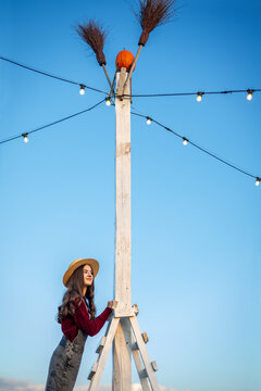 Young woman in straw hat beside a tall wooden pole with pumpkin and brooms, glowing string lights create a festive Halloween scene against clear blue sky.