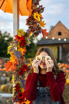 Young woman playfully covering her eyes with two small white pumpkins in autumn garden, colorful seasonal decorations create a cheerful harvest scene.