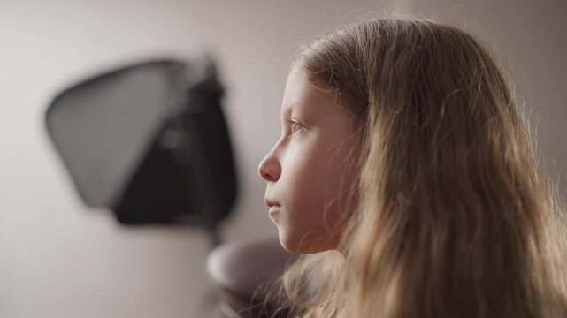 Close up of young girl with long hair in profile view seated indoors under soft lighting. black studio light is visible on wall behind her. scene maintains consistent focus and composition