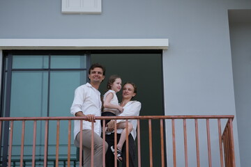 Obraz premium Happy young family with little daughter greet the neighborhood from the balcony with their daughter on the balcony of their new house overlooking the residential estate, candid real moment