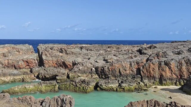 Natural turquoise tidal pool surrounded by rugged coastal rocks with deep blue ocean and clear tropical sky in the background