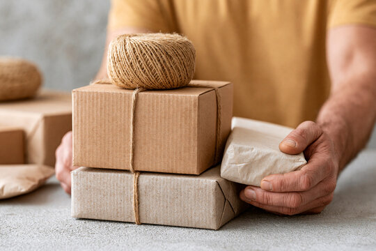 Person holds a wrapped package gently. Brown paper boxes sit stacked neatly. Twine ball rests atop the pile. The hands show care and attention. Ideal for delivery or packaging themes