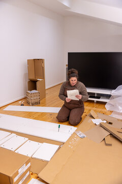 A woman kneeling on a wooden floor reading an instruction sheet while assembling flat‑pack furniture