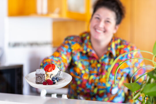 Plate of lamingtons topped with cream and fresh berries held by smiling woman in kitchen