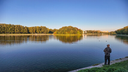 Fototapeta premium A person is fishing at a lake in a park. The scene shows calm water and trees around the lake. It is a bright day with clear skies and no clouds.
