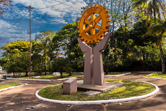 A public monument featuring the iconic golden wheel symbol of Rotary International, held by a concrete sculpture of hands in a green square park 