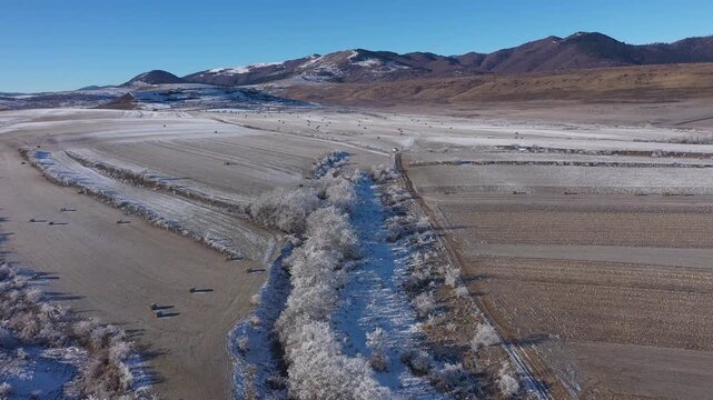  Aerial view of snow-covered field dotted with hay bales under a clear blue sky in a mountainous region during winter