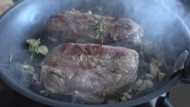 Cooking steaks, close-up. Searing steaks until a golden-brown crust forms. A person uses tongs to flip the steaks in a pan with garlic and herbs.