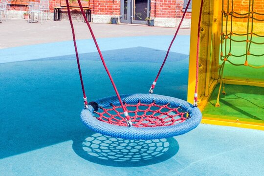 Colorful circular swing with red netting and blue ropes hangs in a sunlit playground, casting a shadow on the blue rubber floor.