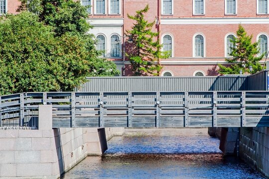 Bridge over the canal at New Holland Island in Saint Petersburg, framed by greenery and a historic brick building with arched windows
