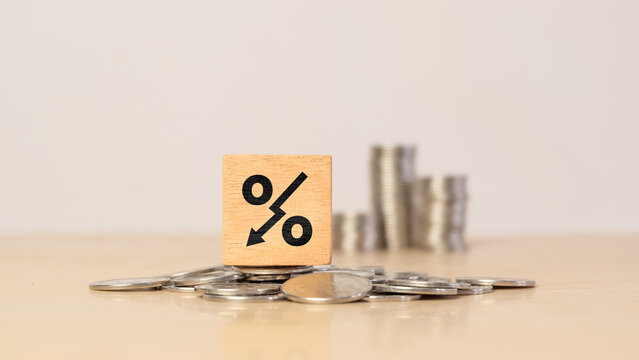 Decreasing percentage and interest rate concept. Wooden cube with falling percent symbol on pile of coins, representing lower rates, financial decline, reduced returns, savings and economic change.