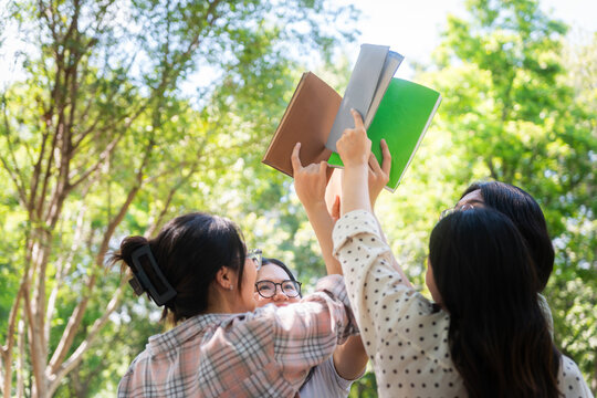 Group of asian university students raising books together in campus park, young students showing teamwork and friendship by holding up books under trees