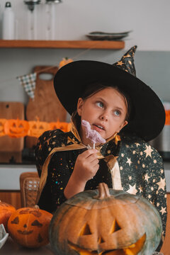 A portrait of a girl in a witch costume eating gummy candies at a Halloween party table. Next to her sits a carved Jack lantern. A children's party during the spooky season. Trick or treat.