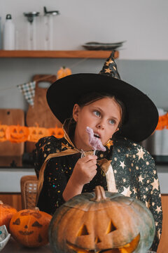 A portrait of a girl in a witch costume eating gummy candies at a Halloween party table. Next to her sits a carved Jack lantern. A children's party during the spooky season. Trick or treat.