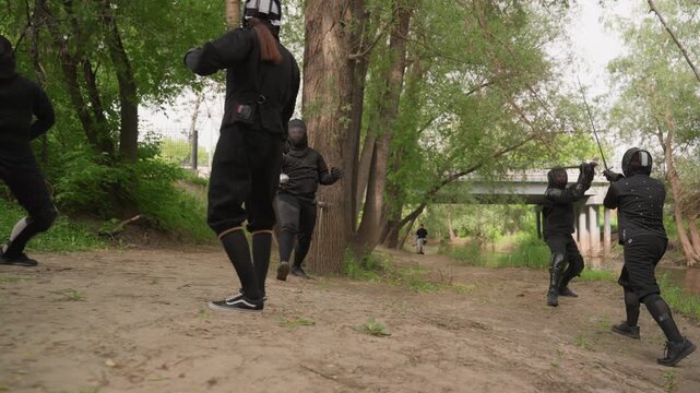 Multiple people in black protective gear engage in sword fighting outdoors along dirt path surrounded by trees and greenery. participants wield swords demonstrating various stances