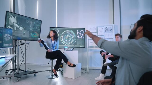 Female engineer presenting a robotic prosthetic arm while explaining robotics technology and digital data to colleagues in a modern laboratory.
