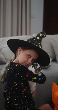 A portrait of a girl in a witch costume celebrating Halloween and hugging her pet rabbit. She is hugging the rabbit and smiling. Celebrating Halloween with children. Trick or treat.