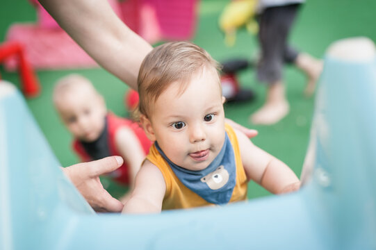 In a lively play zone, a young child climbs on a bright blue structure with joy and wonder, while another baby peeks in the background.