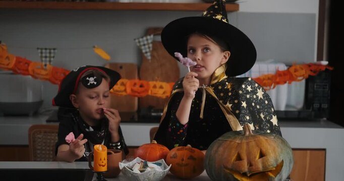 A medium shot of children in Halloween costumes decorating their home, carving a pumpkin, and eating gummy candies. They are playing together and celebrating Halloween in the kitchen. Trick or treat.
