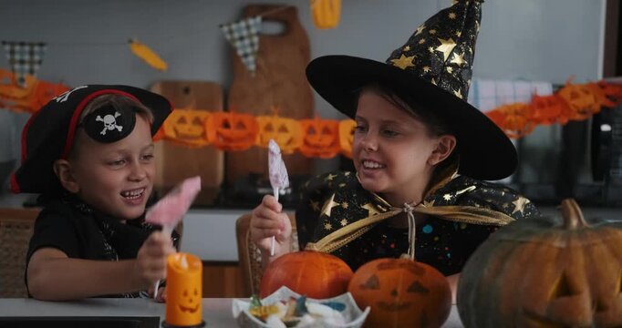 A medium shot of children in Halloween costumes decorating their home, carving a pumpkin, and eating gummy candies. They are playing together and celebrating Halloween in the kitchen. Trick or treat.