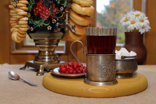 On the table, against the backdrop of the autumn landscape outside the window, there is a glass of black tea in a cup holder and a tray with sugar and sweets, and next to it is a samovar with bagels. 