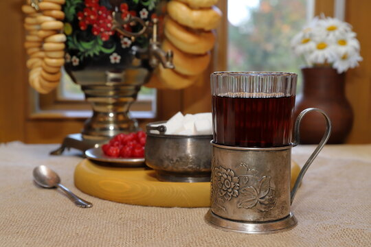 On the table, against the backdrop of the autumn landscape outside the window, there is a glass of black tea in a cup holder and a tray with sugar and sweets, and next to it is a samovar with bagels. 
