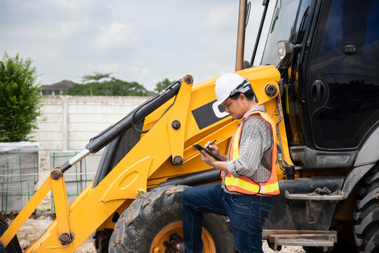 A construction worker white hard hat reflective vest uses tablet stylus beside yellow backhoe loader at work site