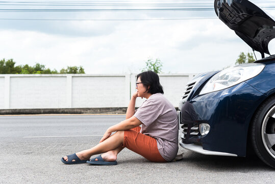 A woman sits the roadside beside blue car its hood open while waiting for help vehicle breakdown