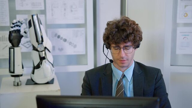 Engineer wearing a headset and working at a computer beside a humanoid robotic hand prototype in a modern robotics research laboratory.
