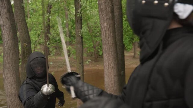 Two women in black fencing gear engage with swords near riverbank demonstrating controlled movements and techniques. Their protective masks obscure faces while surrounding green forest