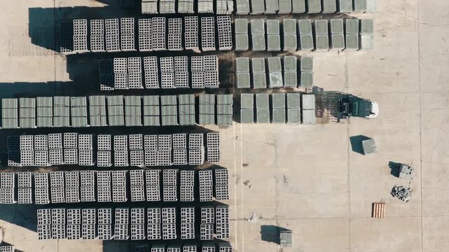Overhead drone footage of a piggyback forklift and flatbed truck working a concrete block storage yard, with long rows of palletized masonry units in hard sunlight.
