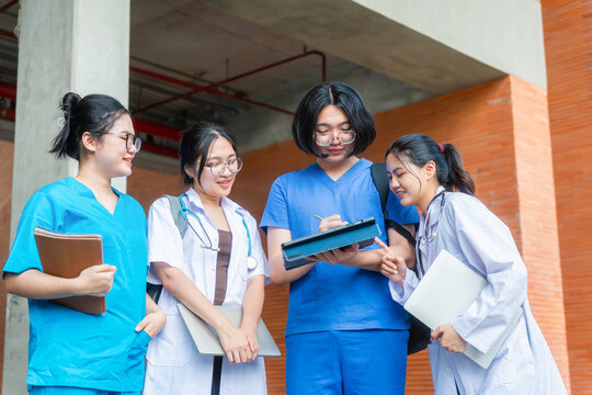 Group of asian medical students or nursing interns discussing with tablet on campus, young healthcare workers in scrubs and lab coats reviewing project outdoors