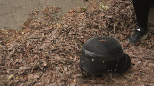 Close up of person in black clothing holding and then dropping old worn fencing mask onto dry leaves on pavement. person is partially visible with focus primarily on helmet descent