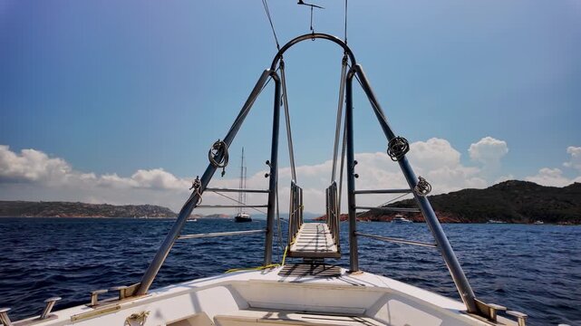 Paniramic view from the bow of a moving cruise ship looking towards the gangway, the open sea, a distant yacht, and a mountainous Mediterranean coastline under a blue, slightly cloudy sky