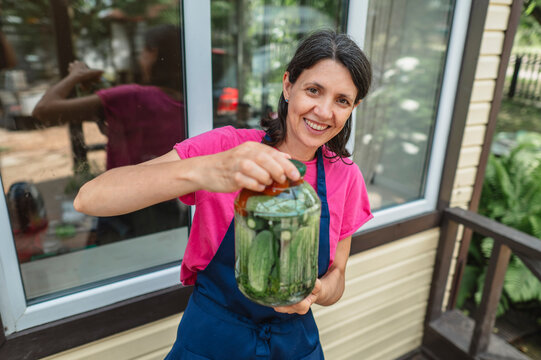 Smiling woman in apron holds glass jar of pickled cucumber outdoors