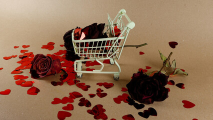 Miniature shopping cart containing dried roses and abundant red heart confetti, symbolizing a past love or the fragility of romantic relationships on a textured brown background © Dark Artistic