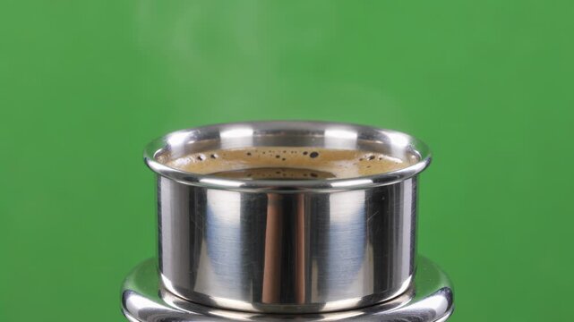 Dark frothy South Indian coffee, mysterious steam swirling from a polished stainless steel dabara set, intensely isolated on  green screen