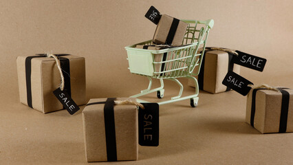Small gift boxes wrapped in craft paper and black ribbons, displaying SALE tags, are placed inside and around a light green shopping cart on a brown background, representing retail offers © Dark Artistic