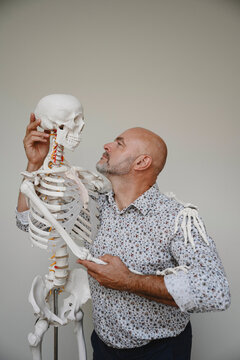 Mature male osteopath holds a medical skeleton model indoors