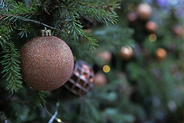 Copper glitter ball hangs among green needles. Wire with tiny lights wraps branch. Dark blurred background with other ornaments. Background for New Year's or Christmas greetings © Alla